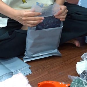 May include: A gray plastic shipping mailer being sealed, containing a dark garment with a subtle pattern. Nearby are stacks of additional gray mailers, several items wrapped in clear plastic, and an orange tape dispenser on a wooden surface.