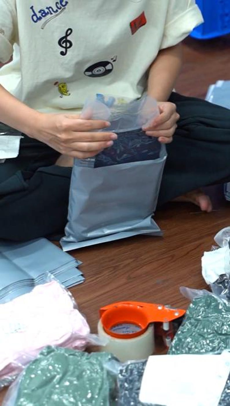 May include: A gray plastic shipping mailer being sealed, containing a dark garment with a subtle pattern. Nearby are stacks of additional gray mailers, several items wrapped in clear plastic, and an orange tape dispenser on a wooden surface.