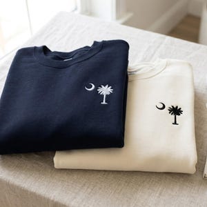 以下が含まれることがあります： 左胸に白い三日月とヤシの木の刺繍が施されたネイビーのクルーネックトレーナーと、同じデザインの黒い刺繍が施されたクリーム色のクルーネックトレーナーが重なっています。どちらもシンプルな長袖のプルオーバータイプです。