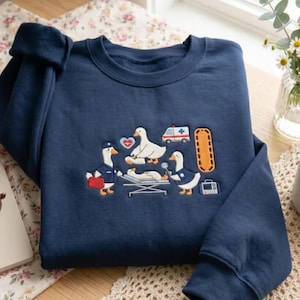 Könnte beinhalten: Ein marineblaues Sweatshirt mit Rundhalsausschnitt und einer Stickerei von weißen Gänsen, die als medizinisches Personal gekleidet sind. Das Motiv zeigt Gänse mit einer Arzttasche, einer Trage, einem Herzsymbol, einem Krankenwagen und einem Monitor.