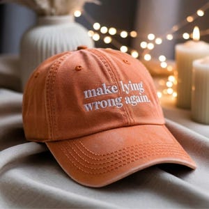 May include: Orange baseball cap with the embroidered text "make lying wrong again." The cap has a curved brim and a fabric strap closure. The hat is made of a soft, textured material.