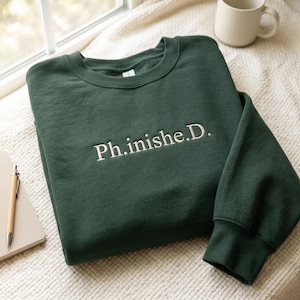 May include: A dark forest green crewneck sweatshirt featuring the embroidered text Ph.inishe.D. in a light cream color across the chest. The garment has long sleeves with ribbed cuffs and a ribbed neckline, presented in a folded, flat-lay orientation.