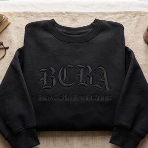 May include: A black crewneck sweatshirt featuring the embroidered text BCBA in a large gothic font, with the words Board Certified Behavior Analyst embroidered in a smaller matching gothic font directly underneath the main lettering.