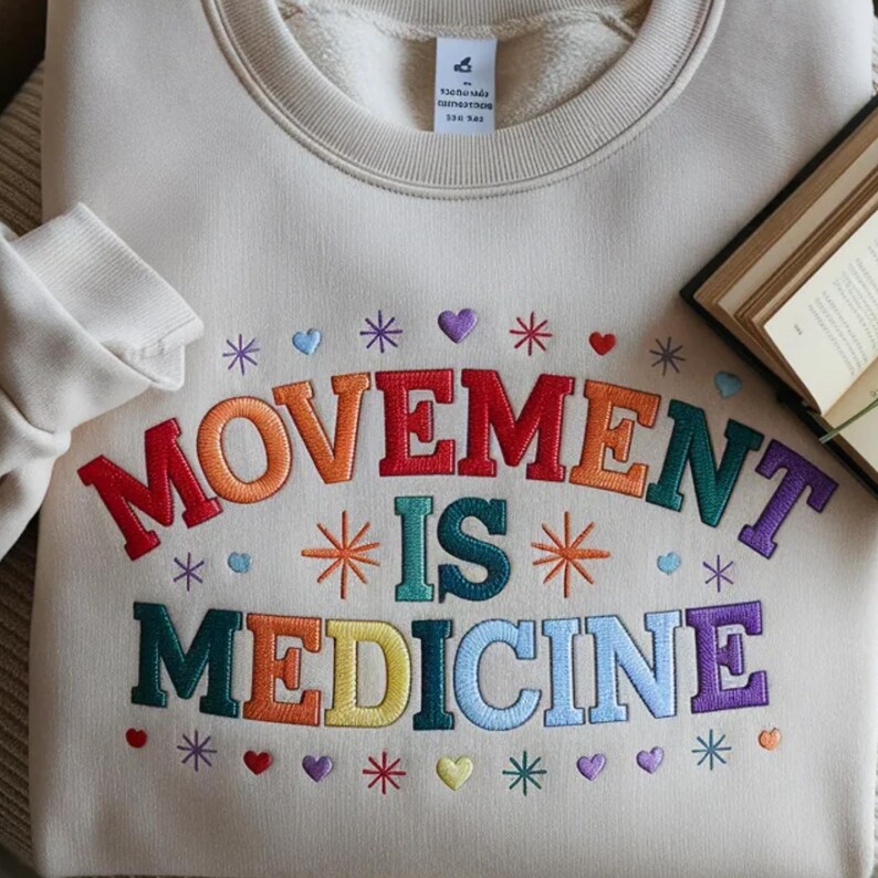 May include: Cream-colored sweatshirt with the embroidered text "MOVEMENT IS MEDICINE" in rainbow colors. The design includes embroidered stars, hearts, and other decorative elements. A book is visible in the background.