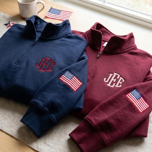 May include: A navy blue and a maroon quarter-zip sweatshirt, each featuring a monogram with the letters JEE embroidered on the chest and a small American flag patch on the sleeve. The navy sweatshirt has red embroidery, while the maroon one has white.