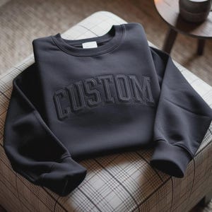 May include: A charcoal grey crewneck sweatshirt featuring the word CUSTOM embroidered in a matching dark thread across the chest in a bold, arched collegiate-style font. The garment has long sleeves with ribbed cuffs and a ribbed neckline.