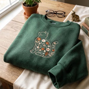 Op de afbeelding: Een donker bosgroene sweater met ronde hals en een geborduurd ontwerp van een zittende kat gevuld met een bloemenpatroon. Het borduurwerk bevat witte, oranje en lichtgroene bloemen en bladeren, omlijnd met een dunne, lichtgekleurde draad.