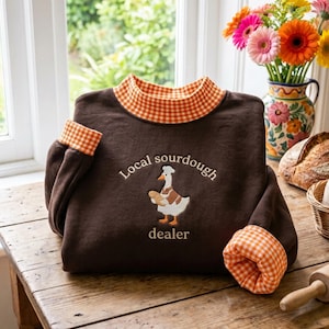May include: A dark brown sweatshirt featuring an orange and white gingham collar and sleeve cuffs. The front displays an embroidered graphic of a white goose wearing a chef hat and holding a loaf of bread, with the text Local sourdough dealer.
