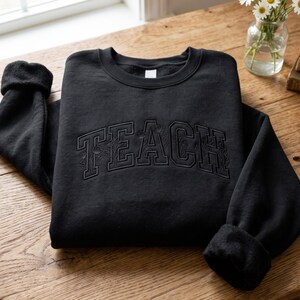 Könnte beinhalten: Ein schwarzes Langarm-Sweatshirt mit dem Wort TEACH, das in einer großen, geschwungenen College-Schrift auf der Brust gestickt ist. Die Buchstaben sind mit schwarzem Garn umrandet und mit einem dezenten, tonalen Blumenmuster gefüllt.
