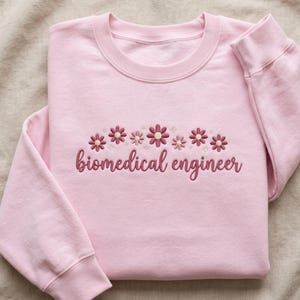 May include: A light pink sweatshirt with embroidered flowers and the words "biomedical engineer" in a matching color. The sweatshirt has a crew neck and long sleeves with ribbed cuffs.