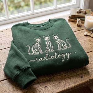 May include: A dark forest green crewneck sweatshirt featuring an embroidered design of three white cat skeletons sitting in a row. Below the cats, the word radiology is written in a white cursive script font. The garment has long sleeves.