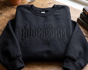May include: A black crewneck sweatshirt featuring the word BOOKWORM embroidered in a dark, gothic-style font across the chest. The garment has long sleeves, a ribbed neckline, and ribbed cuffs, showcasing a monochromatic design with tonal stitching.