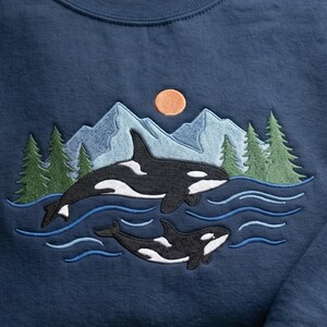 May include: A navy blue sweatshirt featuring an embroidered design of two black and white orcas swimming in blue water. The scene includes light blue mountain peaks, green pine trees, and a small orange sun in the background above the whales.