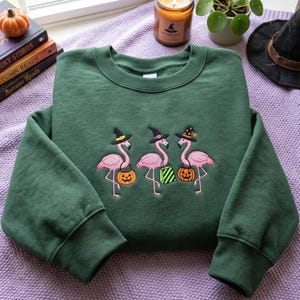 Pode incluir: Uma camisola de gola redonda verde-escura com um bordado de três flamingos cor-de-rosa a usar chapéus de bruxa pretos. Os flamingos transportam artigos de Halloween, incluindo dois baldes de abóbora cor de laranja e um saco às riscas verdes.