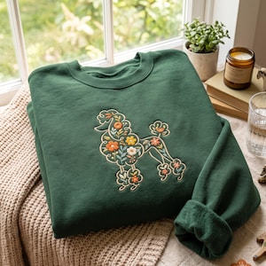 May include: A forest green crewneck sweatshirt featuring a central embroidered design of a poodle silhouette filled with colourful floral patterns including orange, yellow, and white flowers with green leaves and stems on the chest area.