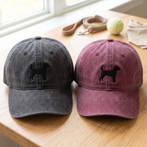 May include: Two baseball caps, one in a washed charcoal grey and one in a washed burgundy red, each featuring a black embroidered silhouette of a dog on the front panel. Both caps have a structured crown, a curved brim, and stitched eyelets.