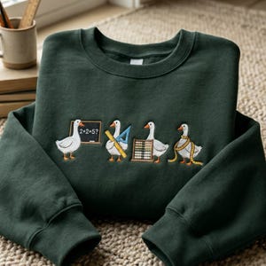 Può includere: Una felpa girocollo verde bosco scuro con un ricamo di quattro oche bianche impegnate in attività scolastiche. Un'oca è accanto a una lavagna con scritto 2+2=5, un'altra tiene un righello giallo e una squadra blu, la terza usa un pallottoliere e la quarta indossa un metro a nastro giallo.
