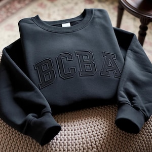 May include: A black crewneck sweatshirt with the embroidered letters "BCBA" across the chest. The sweatshirt has ribbed cuffs and a ribbed hem. The garment is folded on a patterned surface.