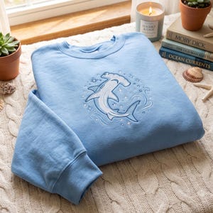 Könnte beinhalten: Ein hellblaues Sweatshirt mit Rundhalsausschnitt und einer weißen Stickerei eines Hammerhais, der von kleinen Sternen und wirbelnden Wellenmustern umgeben ist. Die Stickerei befindet sich mittig auf der Brust des langärmeligen Kleidungsstücks mit Rippbündchen.
