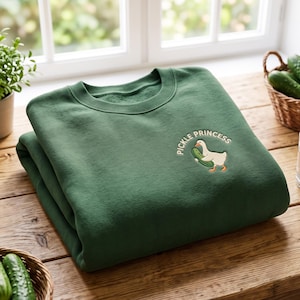 Puede incluir: Una sudadera de cuello redondo color verde bosque con un bordado en el pecho izquierdo. El diseño muestra un ganso blanco con patas naranjas sosteniendo un pepinillo verde, junto al texto arqueado "PICKLE PRINCESS" en letras blancas.