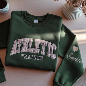 May include: A dark green sweatshirt with the words "ATHLETIC TRAINER" embroidered in a light tan color across the chest. The sleeve features a heart and the name "Sophia" embroidered in a script font.