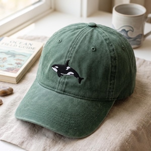 以下が含まれることがあります： フロントパネルの中央に黒と白のシャチの刺繍が施された、フォレストグリーンのベースボールキャップ。このキャップは、構造化されたクラウン、カーブしたつば、通気用のステッチ入りアイレットを備えており、コットン生地で作られています。