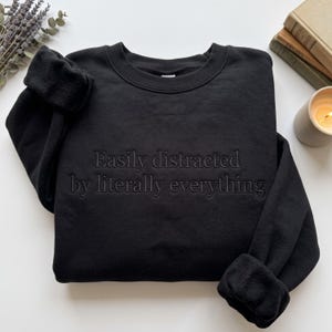 May include: A black crewneck sweatshirt featuring the embroidered text "Easily distracted by literally everything" in a matching black thread. The garment has long sleeves with ribbed cuffs and a ribbed neckline, creating a monochromatic design.