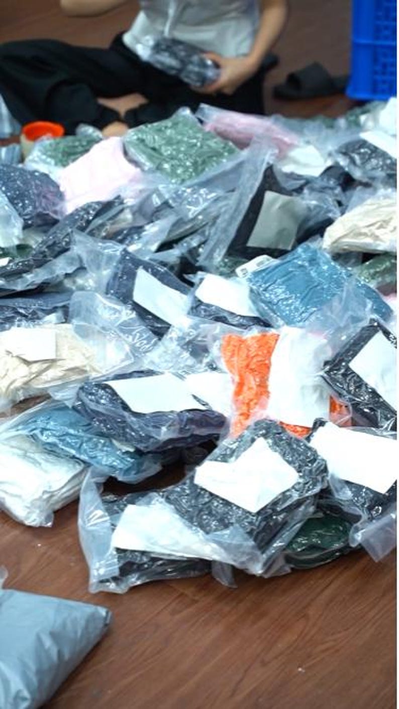 May include: A large collection of assorted apparel items individually packaged in clear plastic bags. Each bag features a white rectangular label on the front. The items vary in color, including black, white, orange, green, and beige fabric.