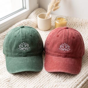 May include: Two baseball caps, one in a muted forest green and the other in a faded brick red, each featuring a white embroidered lotus flower design centered on the front panel. Both caps have a structured crown, a curved brim, and eyelets.