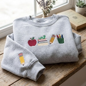May include: A light gray crewneck sweatshirt featuring embroidered school-themed designs. The chest displays a red apple, a stack of books with a green worm, a yellow pencil, and a green cup with crayons. A single yellow pencil is embroidered on the sleeve.