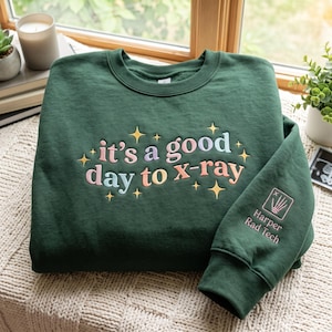 以下が含まれることがあります： パステルカラーで「it's a good day to x-ray」という刺繍文字と小さな黄色い星が施されたダークグリーンのクルーネックスウェットシャツ。袖には手の骨格の四角い刺繍アイコンと「Harper Rad Tech」という文字が入っています。