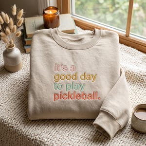 Peut inclure: Un sweat-shirt beige à col rond avec le texte brodé "it's a good day to play pickleball." Les mots sont cousus en format empilé en utilisant quatre couleurs distinctes : du fil rose, jaune, vert clair et orange pour les lettres.