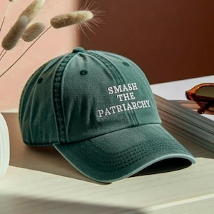 Peut inclure: Casquette de baseball vert foncé avec le texte "SMASH THE PATRIARCHY" en blanc. La casquette a une visière incurvée et une fermeture à sangle en tissu. Le chapeau est posé sur une surface claire.