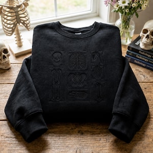 May include: A black crewneck sweatshirt featuring tonal black embroidery of various human skeletal structures, including a skull, rib cage, pelvis, and foot bones, arranged in a grid pattern on the front of the garment with long sleeves.