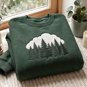 May include: A dark forest green crewneck sweatshirt featuring a central embroidered design of a white textured cloud positioned above a dense cluster of dark green pine trees. The garment has a ribbed collar, cuffs, and hem, with a soft fleece interior.