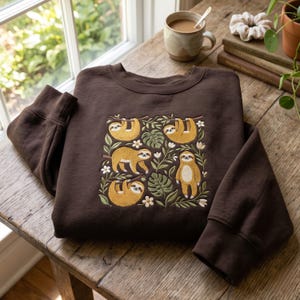 Peut inclure: Un sweat-shirt à col rond marron foncé avec une broderie représentant quatre paresseux suspendus à des branches parmi des feuilles de monstera vertes et de petites fleurs blanches. Le motif est centré sur la poitrine avec des détails.