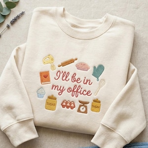 Op de afbeelding: Crèmekleurige sweatshirt met geborduurde bakgerelateerde afbeeldingen. Voorzien van een mixer, deegroller, taart, ovenwant, garde, kookboek, cupcake, suikerpotten, eieren en een weegschaal. De tekst luidt "I'll be in my office" in een sierlijk lettertype.