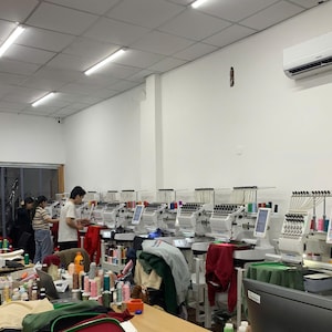 May include: A row of industrial embroidery machines arranged in a bright workshop, featuring multiple needle heads, digital control screens, and various colorful thread spools, with workers operating the equipment to process garments.