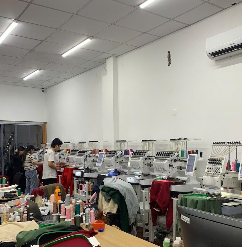 May include: A row of industrial embroidery machines arranged in a bright workshop, featuring multiple needle heads, digital control screens, and various colorful thread spools, with workers operating the equipment to process garments.