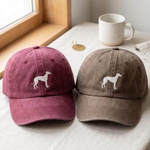 May include: Two baseball caps, one in a deep maroon and one in a muted brown, each featuring a white embroidered silhouette of a slender dog standing in profile. Both caps have a structured crown, a curved brim, and stitched eyelet details.