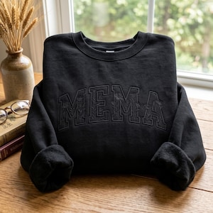 Könnte beinhalten: Ein schwarzes Sweatshirt mit Rundhalsausschnitt und dem Wort MEMA, das in einer großen, geschwungenen Schriftart im College-Stil gestickt ist. Die Buchstaben sind mit einem dezenten, tonalen Blumenmuster gefüllt.