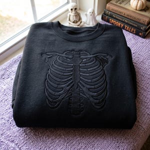 Könnte beinhalten: Ein schwarzes Sweatshirt mit Rundhalsausschnitt und einer tonalen, erhabenen Stickerei eines menschlichen Brustkorbs auf der Vorderseite. Die Stickerei ist schwarz und passt zum Stoff des Sweatshirts, was ein dezentes, strukturiertes Skelettmotiv ergibt.