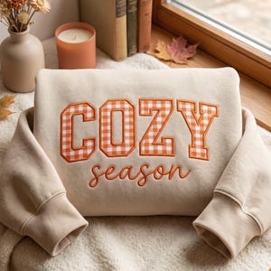 May include: A beige sweatshirt featuring the word COZY in large, orange and white gingham-patterned block letters, with the word season embroidered in orange cursive script underneath. The garment has long sleeves with ribbed cuffs.