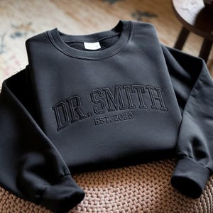 以下が含まれることがあります： 胸元にカレッジスタイルのフォントで「DR. SMITH」および「EST. 2026」と刺繍された黒のクルーネックトレーナー。長袖で袖口と襟元はリブ編みになっており、すっきりとした構造的なデザインが特徴です。