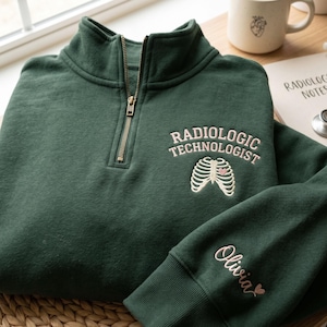 Könnte beinhalten: Dunkelgrünes Sweatshirt mit Viertelreißverschluss und rosa gesticktem Schriftzug "RADIOLOGIC TECHNOLOGIST" über einer Rippenkorb-Grafik. Der Ärmel zeigt den Namen "Olivia" in rosa Schrift mit einem Herz. Eine Tasse und ein Notizblock sind im Hintergrund.