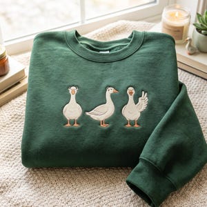 Puede incluir: Una sudadera de cuello redondo en color verde bosque oscuro con un diseño bordado de tres gansos blancos. Los gansos están colocados en fila, con el ganso de la izquierda mirando al frente, el central de perfil y el de la derecha saludando.