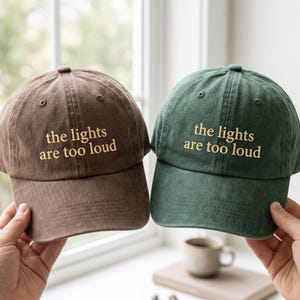 May include: A brown baseball cap and a dark green baseball cap, both featuring the embroidered text "the lights are too loud" in gold thread. Each cap has a six-panel construction with a curved brim and a button on the top center.