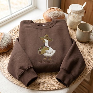 May include: A brown sweatshirt with a whimsical embroidered design. The design features a white goose wearing a sombrero and holding a taco. The text "ME GOOSE-TA" is below the goose.