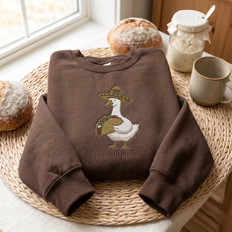 May include: A brown sweatshirt with a whimsical embroidered design. The design features a white goose wearing a sombrero and holding a taco. The text "ME GOOSE-TA" is below the goose.