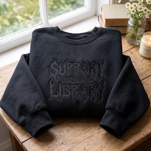 Może przedstawiać: Czarna bluza z okrągłym dekoltem i tonalnym czarnym haftem na piersi. Napis "SUPPORT YOUR LOCAL LIBRARY" wykonano stylizowaną, poszarpaną czcionką. Odzież posiada długie rękawy z prążkowanymi mankietami oraz prążkowany dekolt wykonany z tkaniny.